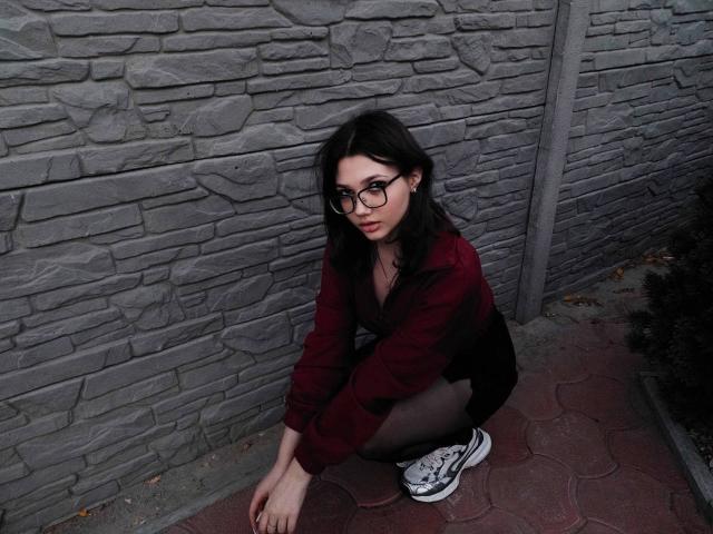 EllieJulse con gli occhiali, accovacciata, indossa una giacca di velluto rosso, una gonna nera e scarpe da ginnastica bianche con dettagli neri, davanti a un muro di pietra grigia. Ha un bel sedere..
