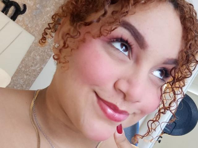 YeraCarey with curly red hair, pink blush, red lipstick, and eye makeup, smiling up, showing nose and upper chest, wearing a necklace, with red nail polish.