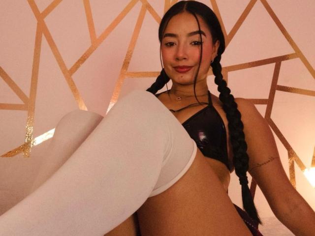 NicoleRivera with braided hair, wearing a black bra, and white thigh-highs, showing her ass while sitting, looking at the camera, with a subtle smile.