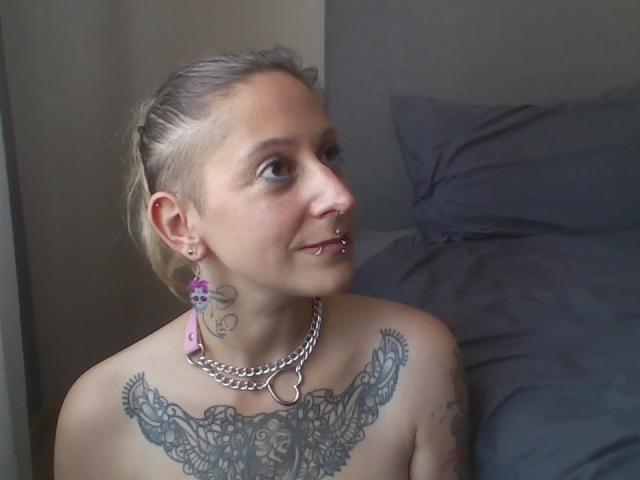 Topless MissLilis with blonde braids, nose ring, and lip piercing, wearing a chain necklace, has a large butterfly tattoo across her tits, and purple earrings.