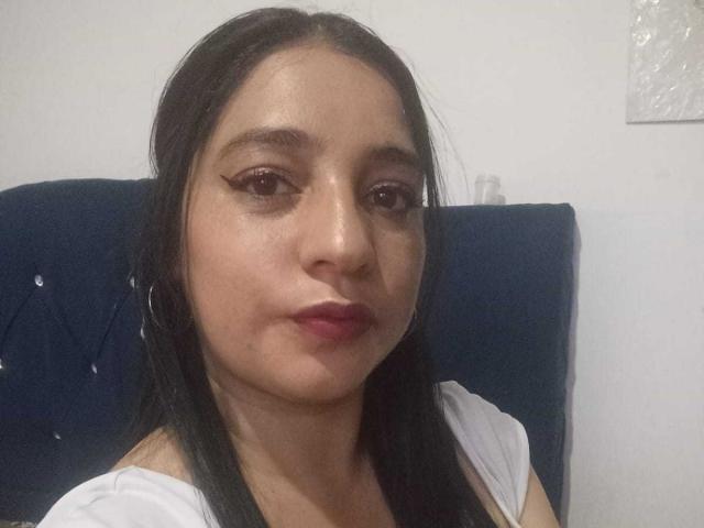 ElizaLowry has medium brown skin, straight black hair, and wears a white top. She has winged eyeliner, pink lipstick, and small hoop earrings, looking slightly pouty.