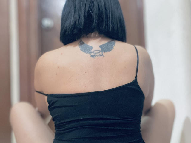 SamantaMuller has black straight hair, a black tattoo with wings and a skull on her back, wearing a black spaghetti strap top, sitting with her back to the camera.