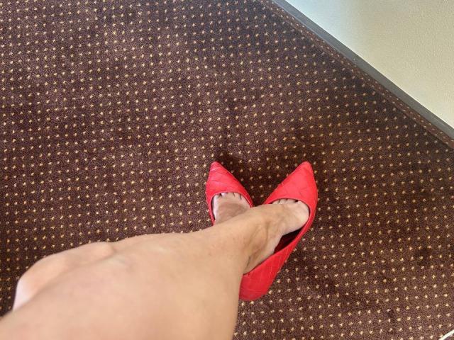MoonLiza's leg in red pointed heels, showing a fair skin tone, with a brown textured carpet. Her foot is slightly pointed, with a visible ankle.