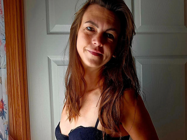 LillyMania-hot with dirty brown hair, smiling, wearing a black lace bra, standing in front of a white door, sunlight on her face, looking cute and slightly sweaty.