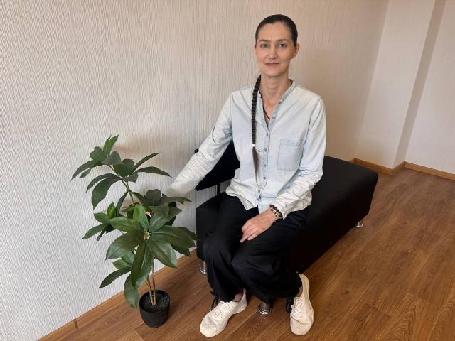 TayaFlavy with braided hair, light blue shirt, black pants, white shoes, sitting on black bench, touching green plant, wooden floor, white wall, small smile, casual.