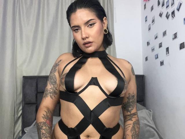 VictoriaVicious with tattoos, black strap harness, and nose ring, showing her tits, in a bedroom. She has dark hair, hoop earrings, and a confident expression.