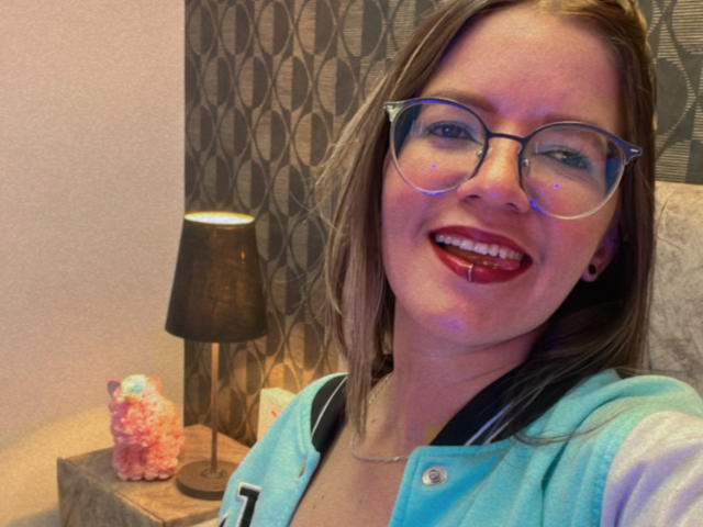 AliceHoe met een bril, rode lippenstift en bloedige tanden, draagt een blauwe hoodie, glimlacht op een selfie, heeft een kleine neus, lichte huid en bruin haar..
