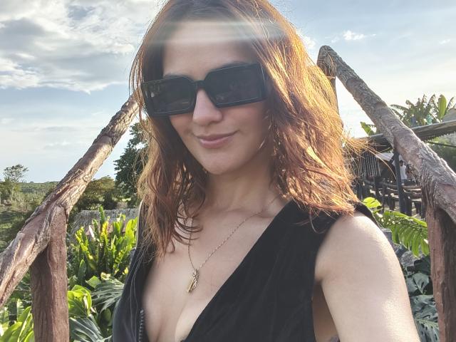 AmelieLautrec has wavy brown hair, wears black sunglasses, and a low-cut black top showing her tits. Sunlight beams through tree branches, highlighting her smile.