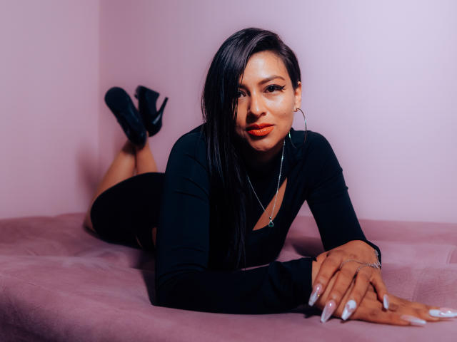 ZaiiraFox sdraiata su un letto rosa, indossa una maglia nera a maniche lunghe e tacchi neri, con i glutei in alto e le mani davanti, ha lunghi capelli neri e rossetto rosso..