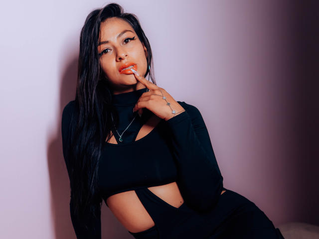 ZaiiraFox, com longos cabelos pretos, vestindo uma blusa preta com recortes, com o dedo na boca, olhando para a câmera, fundo rosa suave, sexy, atraente, boca ligeiramente aberta, mostrando batom vermelho..
