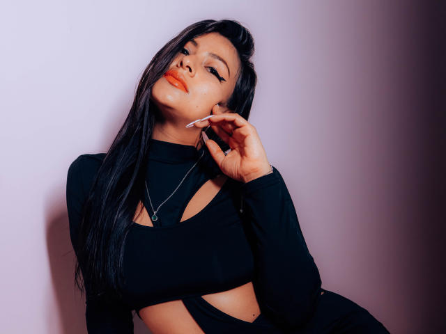 ZaiiraFox met lang zwart haar, die een zwarte top met uitsparingen draagt, een sigaret bij haar mond houdt, naar beneden kijkt met oranje lippenstift, en een zilveren ketting draagt..