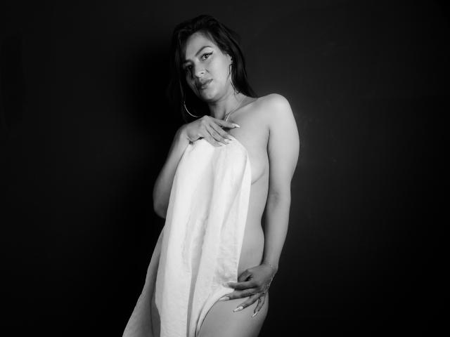 Zwart-wit foto van ZaiiraFox met een gemiddelde borstmaat, die een handdoek vasthoudt om haar billen te bedekken, staand voor een donkere achtergrond, met oorringen, en kijkend naar de camera..