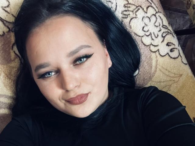 MaxieNelly has pale skin, black eyeliner, and dark lipstick. She's lying down with black hair, wearing a black top, and has a subtle smile. Her tits are not visible.