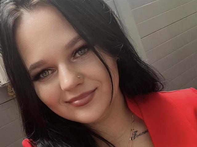 MaxieNelly has long black hair, green eyes, a small nose piercing, and a subtle smile, wearing a red top with a necklace, looking cute and sexy.