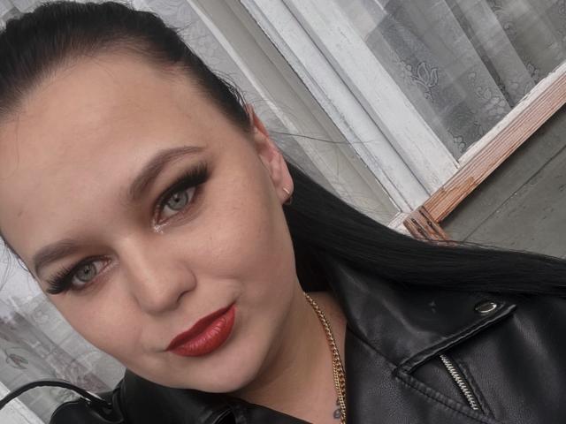 MaxieNelly with pale skin, black hair in a ponytail, red lipstick, black eyeliner, and a black leather jacket, wearing a gold chain necklace.
