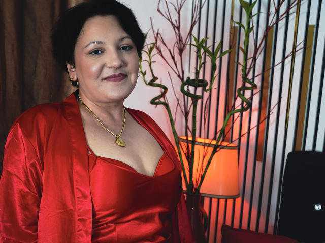 MayaBlowsYou, une femme d'âge moyen avec des cheveux noirs courts, porte une robe en satin rouge avec un décolleté bas, qui révèle ses petites seins, et un collier en or, tout en souriant..