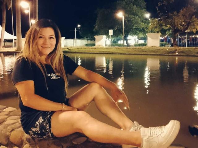 DanaWester with long blonde hair, black t-shirt, and white sneakers, sitting on rocks by a reflective waterway at night, smiling, wearing floral shorts, legs crossed.