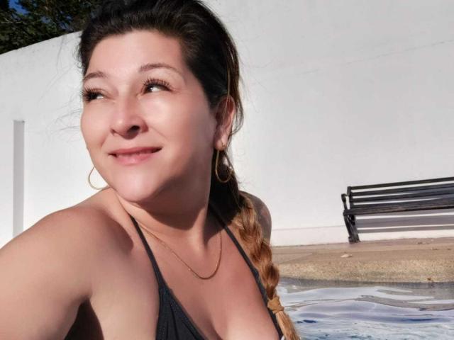 DanaWester with a smile, brown braid, black bikini top, showing her boobs, gold hoop earrings, and a gold necklace, standing by a pool.