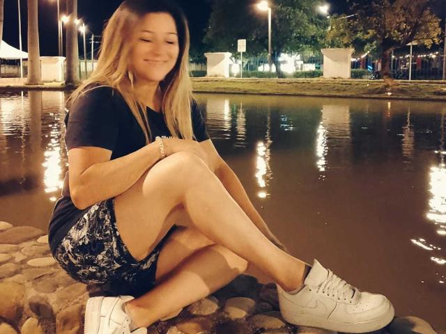 DanaWester with long blonde hair, smiling, sitting on rocks by a reflective waterway at night. Wearing a black shirt, floral shorts, and white sneakers. Hands on knees.