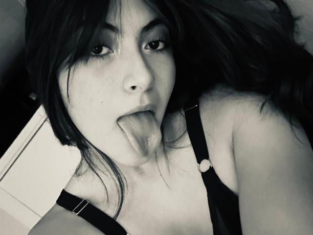 SpencerLe with dark hair, sticking out her tongue, wearing black straps, has a nose ring, and her tits are partially visible in a black-and-white selfie.