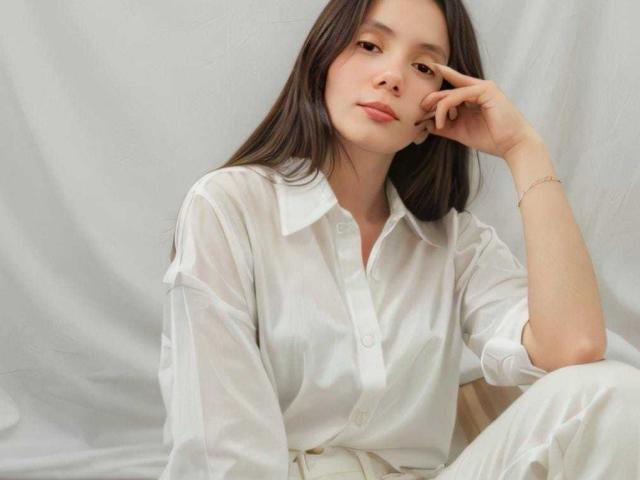 HallieMils with long brown hair, wearing a sheer white button-up shirt, sitting with a relaxed, slightly bored expression, hand touching her cheek, wearing a simple gold bracelet.