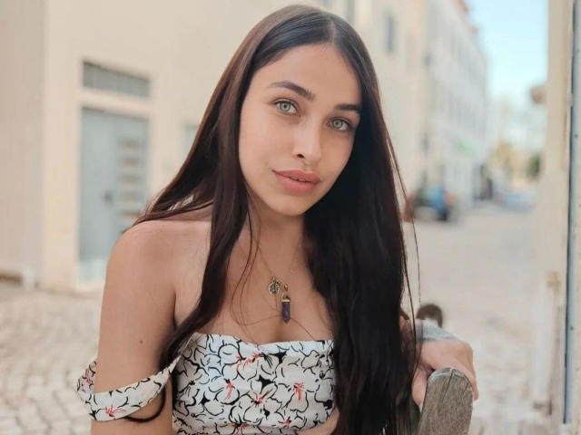 NicoleJules-hot with long dark hair, green eyes, and a floral off-shoulder top, showing her small tits and a necklace with a purple stone, in a sunlit street.