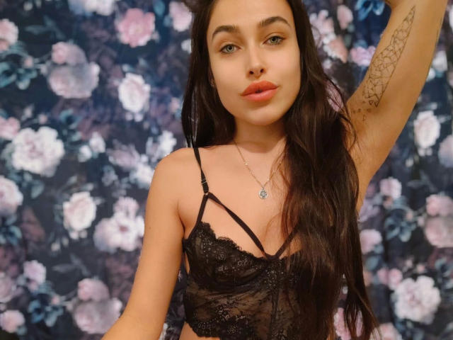 NicoleJules-hot with long dark hair, wearing a black lace bra, pouty lips, arm tattoo, and necklace, looking at camera, holding arm up, showing small tits.