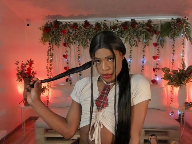VenusMiller with long black pigtails, holding a pigtail, wearing a white crop top with red plaid bow, red lips, in a red-lit room, looking at the camera.
