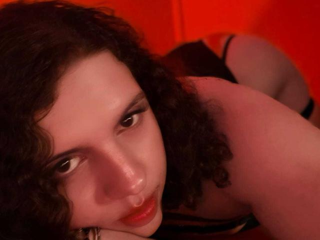 CatleyaNoire mit lockigem, dunklem Haar, blasser Haut und roten Lippen, liegt auf der Seite, blickt mit halb geschlossenen Augen nach oben und trägt schwarze Dessous, wobei ihr Hintern leicht angehoben ist..