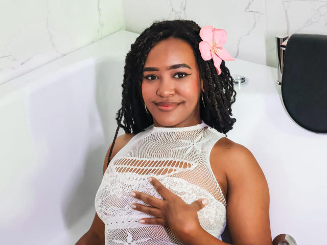 NaomiReyes with dark curly hair, pink flower, and white mesh top, touching her tits in a bathtub, smiling. She has a cute face and brown skin.