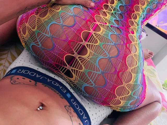 Le bas en dentelle coloré de FretcyAmber recouvre ses fesses, elle porte des sous-vêtements blancs à pois avec une ceinture "BOYNYORK", on peut voir ses tatouages, son piercing au nombril, et elle a les mains sur ses fesses..