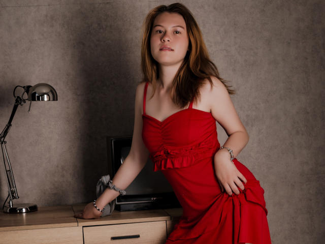 TaylorCarpert with light brown hair, wearing a red dress, has her hands cuffed to a wooden table, leaning forward, and looking at the camera. Her tits are visible.
