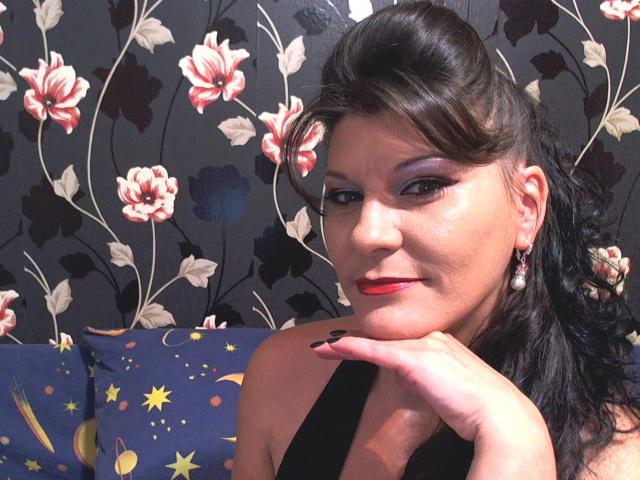 Emmahotcum has dark hair in a retro updo, purple eyeshadow, red lipstick, black dress, and pearl earrings, resting her chin on her hand with black nails.