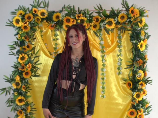 SofiFw with long red braids, black top, and shiny black skirt, standing in front of a sunflower and green leafy wreath, yellow fabric backdrop, smiling.