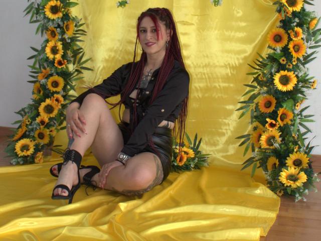 SofiFw with long red braids, black jacket, and shorts, sitting on yellow satin, surrounded by sunflowers, wearing black heels, showing her ass and tattoos, smiling.