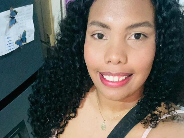 ElizabethMona, with curly black hair, smiling, pink lipstick, brown eyes, and a necklace, wearing a black strap top, has a cute face and a slightly chubby, sexy ass.