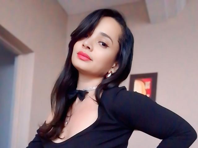 LucyDelia with black hair, red lipstick, black bow choker, black top, small tits, light skin, earrings, slightly tilted head, confident expression, soft lighting, indoor setting, relaxed pose.