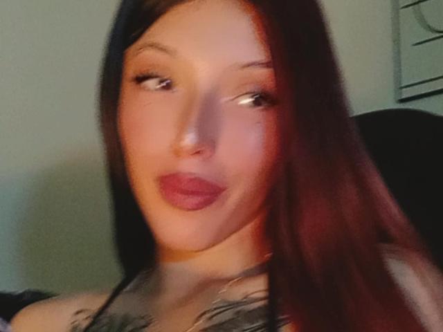AizenMyoo with long brown hair, light skin, and red lipstick, looking to the side with a slight smile. Her tits are partially visible with a tattoo on her chest.