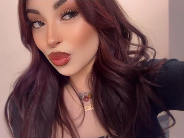 MaddyCalloway has dark red, wavy hair, full red lips, and a sexy, confident face. She's wearing a black top and a necklace with colorful beads.