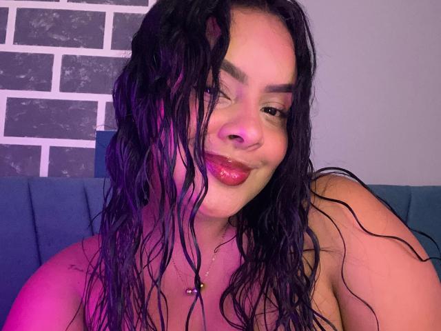 AmarantaCarey with wet black hair, purple-lit skin, and shiny red lips, smiling at the camera, showing a nipple piercing. Boobs visible. Casual, confident.