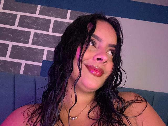 AmarantaCarey has wet black hair, pink lipstick, and a shiny nose, smiling with pink highlights on her cheek, wearing a necklace, sitting against a blue and grey wall.