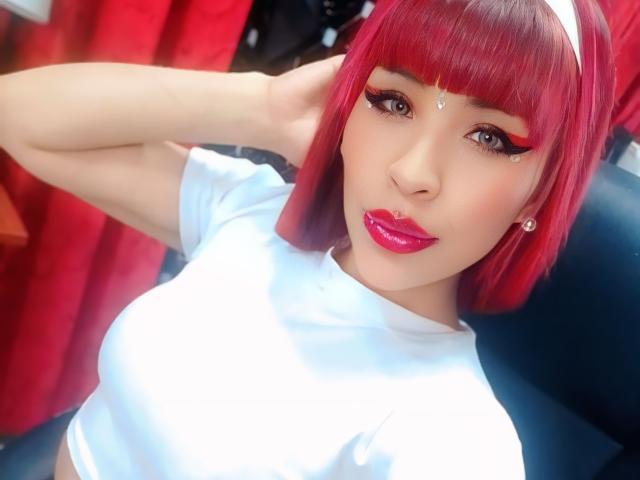 MilleiGrey with red bob, white headband, and white tee, has shiny pink lips, eyeliner, and a jewel on her forehead. Her hands are behind her head, showing her tits.