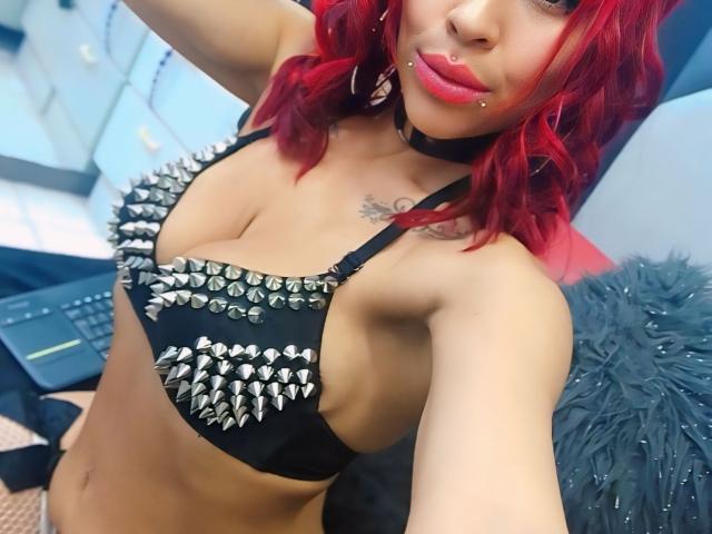 MilleiGrey with red hair, black spiked bra, choker, and lip piercing, showing her tits, tattoos, and pink lipstick, in a selfie. Laptop and gray fur pillow in background.