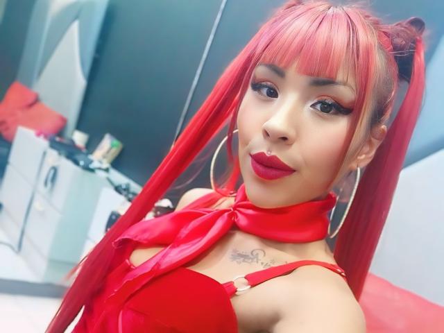 MilleiGrey mit roten Zöpfen, rotem Lippenstift, großen Ohrringen, einem roten Schal und einem roten Kleid, die Tattoos auf der Brust zeigt, lächelnd, in einem Selfie. Brüste sind unter dem Kleid sichtbar..