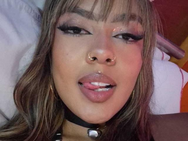 AmelieDuvall with brown hair, nose rings, and a black choker, tongue out, glossy pink lips, and dramatic eyeliner, looking at the camera. Her lips are slightly parted.