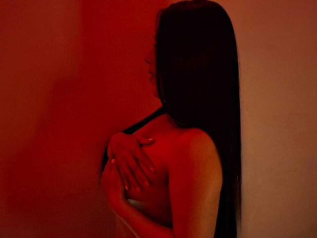 SylvieColette mit langem, schwarzem Haar, in rotem Licht, bedeckt ihre nackten Brüste mit einer Hand, steht an einer Wand, blickt zur Seite, in einem sinnlichen, dunklen Foto..