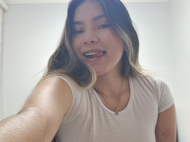 ValeryThomss with long brown hair, tongue out, wearing a white tee, gold necklace, and a small tattoo on her right arm, in a selfie.