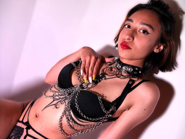CamiVargas with short brown hair, red lipstick, wearing a black bra and strappy lingerie, chains and a spiked collar, yellow nails, looking at the camera, showing her tits.