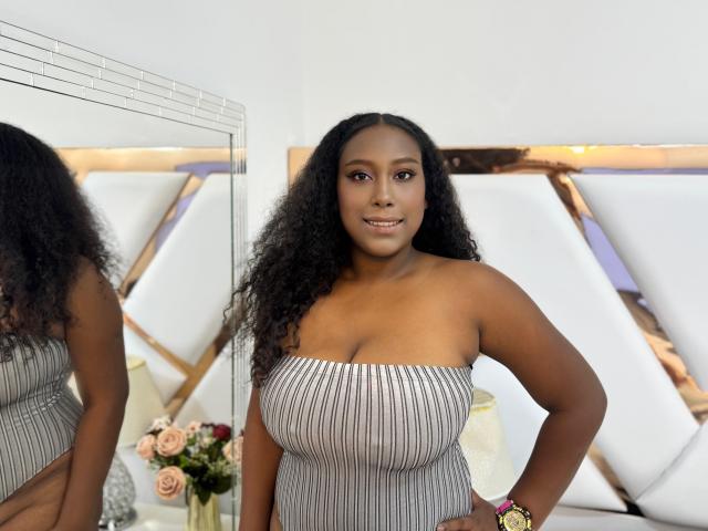 RebecaSolis stands in a room, wearing a strapless, ribbed grey dress, showing her big boobs, dark curly hair, and a small smile. A mirror reflects her on the left.