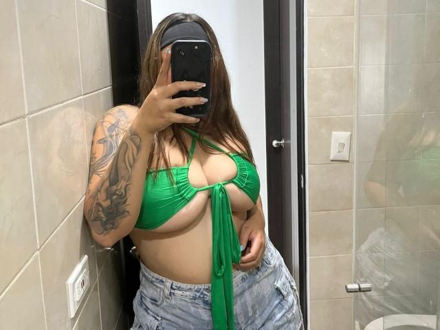 AllisonJustingb in een groen bikini topje, waarbij haar grote borsten zichtbaar zijn, met een getatoeëerde arm, en met een spijkerbroekje, maakt een spiegelzelfportret met een telefoon, in een betegelde badkamer..